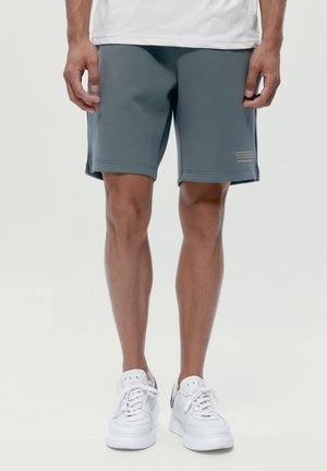 Mann trägt graue kniehohe Shorts, weiße Turnschuhe und ein weißes T-Shirt, steht vor einem schlichten hellen Hintergrund.