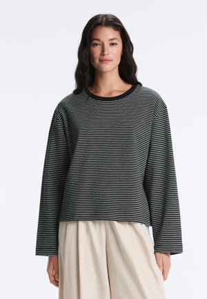 Femeie cu păr lung și închis la culoare, purtând o bluză cu mâneci lungi, cu dungi negre și gri, și pantaloni largi de culoare bej deschis, stând în fața unui fundal alb.