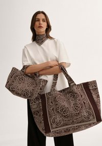 Kobieta w białej bluzce i czarnych spodniach trzyma dwie duże brązowe torby w wzór paisley, po jednej na każdym ramieniu, z pasującą chustą na szyi.