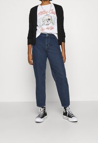 Černý kardigan, bílý tričko s grafikou lebky, modré džíny s volným střihem a černé vysoké tenisky.