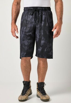 Uomo che indossa pantaloncini sportivi neri con motivo e coulisse e sneaker alte color beige, in piedi davanti a uno sfondo semplice.