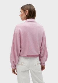 Ružičasti pleteni pulover s ležernim krojem, rebrastim rubom, velikim rukavima i ovratnikom. Kombiniran s bijelim hlačama koje imaju dekorativne detalje u obliku srca.