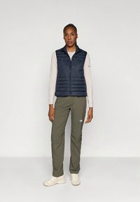 Marineblauw gewatteerd vest met een ritssluiting aan de voorkant, gecombineerd met een licht langärmelig shirt en olijfgroene broek. Model draagt beige schoenen en staat tegen een effen achtergrond.