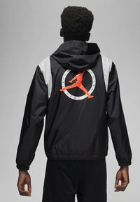 Giacca a vento nera con cappuccio, con dettagli grigi e un prominente logo arancione di Air Jordan sul retro, polsini e orlo elasticizzati.