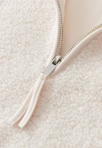 Fleece-Stoff in hellbeige mit einem glatten Reißverschluss. Der Reißverschluss hat einen silbernen Ziehgriff und Metallzähne, die im Kontrast zur weichen Textur stehen.
