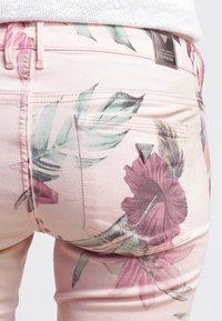 Roze spijkerbroek met grote hibiscus- en bladpatronen. Voorzien van een metalen merkembleem nabij de tailleband en de achterzakken.