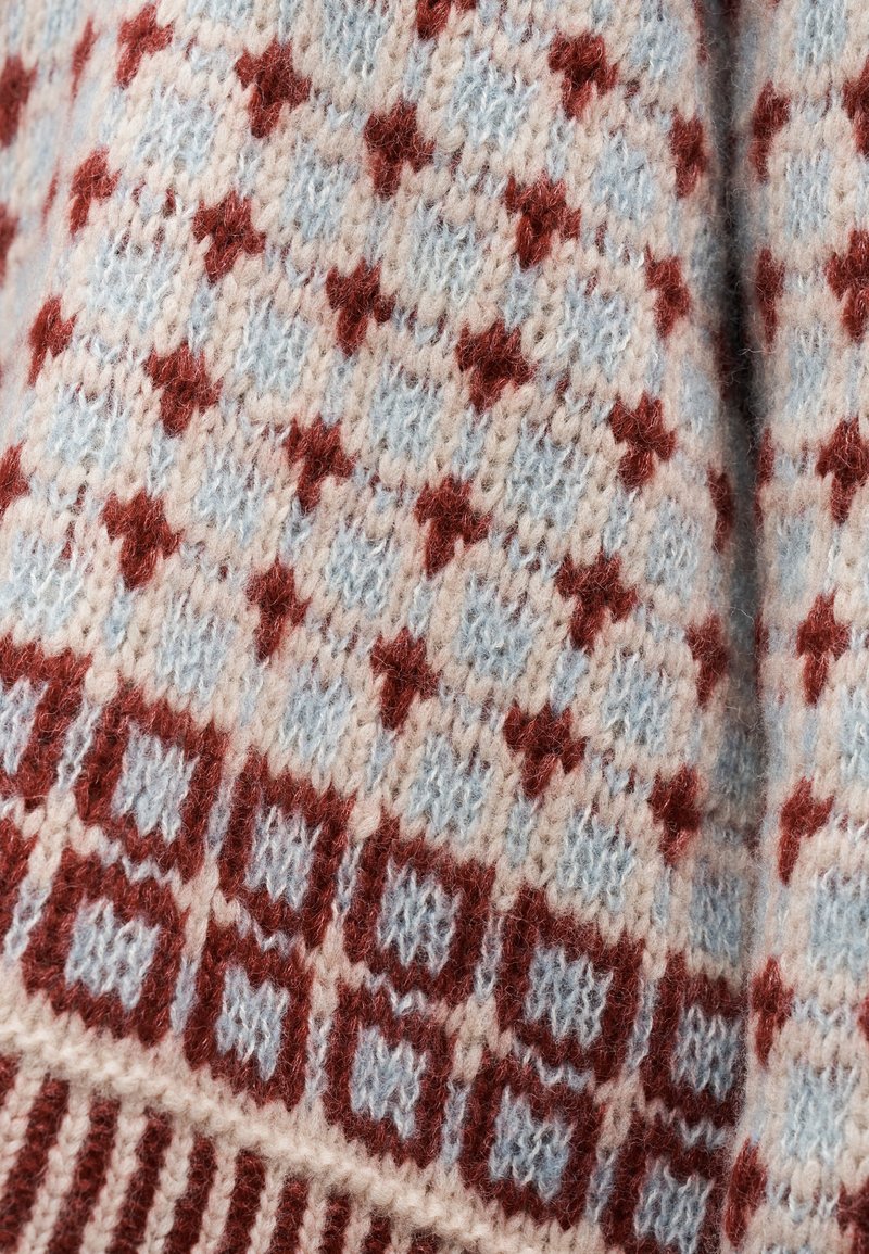 Nahaufnahme eines gestrickten Stoffes mit sich wiederholendem Rautenmuster in Rot, Hellblau und Cremefarben auf einem Pullover.