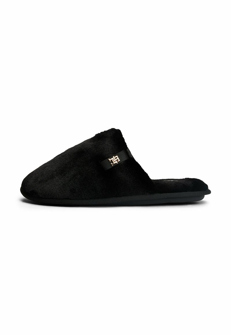 Zapatilla de casa negra de felpa, con una textura suave, diseño de punta redondeada y suela de caucho negra. Presenta un pequeño logotipo en el lateral.