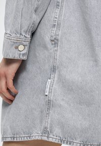 Denimkjole i lysegrå med knappet manchet, sideslids og en mærkat nær kanten. Har en glat tekstur og struktureret design.