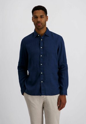 Mann steht und schaut nach vorne, trägt ein navyblaues, langärmeliges Hemd mit Knöpfen und beigen Hosen vor einem einfarbigen hellen Hintergrund.