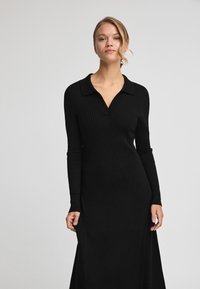 Schwarzes, geripptes Strickkleid mit einem Kragenhals, langen Ärmeln und seitlichen Schlitzen. Hat eine figurbetonte Silhouette und strukturierten Stoff.