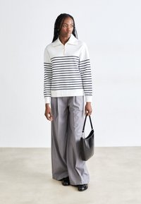 Maglione a righe bianco e navy con colletto a zip, abbinato a pantaloni grigi a gamba larga e una borsa nera. Tessuto morbido e vestibilità rilassata.
