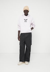 Ροζ παρκά με μαύρο γραφικό σχέδιο, μπροστινή τσέπη, συνδυασμένο με σκούρα γκρι cargo παντελόνι και λευκά αθλητικά παπούτσια. Ουδέτερο υφασμάτινο καπέλο.