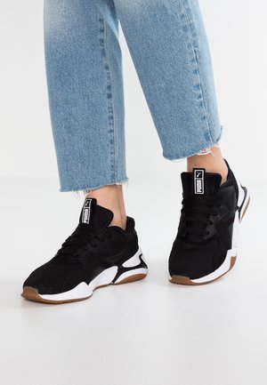 Person iført sorte og hvide Puma-sneakers med gummisåler og lyseblå cropped jeans stående på et hvidt gulv.