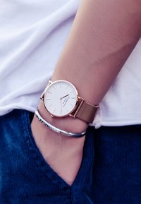 Orologio con cinturino in maglia rosè e quadrante minimalista bianco, caratterizzato da sottili indicatori delle ore e da una cassa elegante. Indossato su un polso con un bracciale argentato.