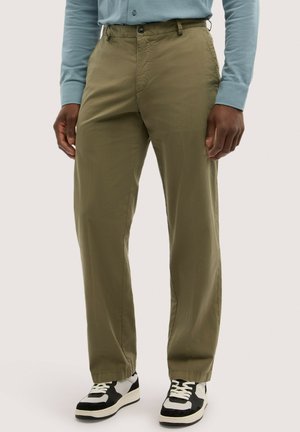 Mand iført olivengrønne bukser med lige ben, lyseblå skjorte med lange ærmer og sort-hvide sneakers, stående mod en ensfarvet baggrund.