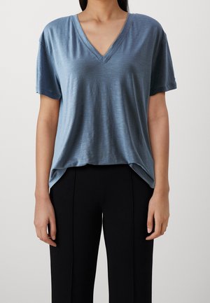 Camiseta azul de cuello en V en tela ligera y suave. Mangas cortas, corte relajado y un poco más larga en la parte de atrás. Combinada con pantalones negros.