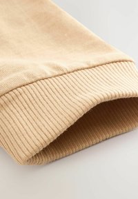 Beige kötött anyag, bordázott mandzsetta részlettel. A felület puhanak tűnik, enyhe színváltozásokkal, az élén hajtva.