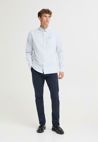 Lyseblåstripet skjorte med knapper og en liten lomme, kombinert med mørk marineblå bukser og svarte sko, båret av en stående ung mann.