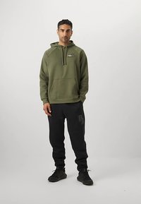 Hoodie verde oliva con tasca frontale e coulisse, abbinato a pantaloni della tuta neri con logo. Sneakers nere completano l'outfit.
