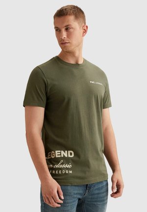 Muž v olivovo zelenej tričku PME Legend s bielym potlačeným textom, v modrých džínsach, stojaci pred jednoduchým svetlým pozadím.