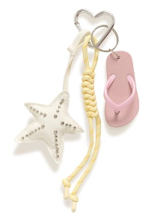 Lyshåret kvinde iført hvid ærmeløs top og cremefarvede bukser holder en vævet tan tote-taske med pink og søstjerne-nøgleringe.