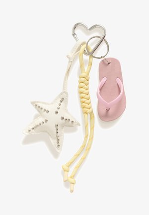 Lyshåret kvinde iført hvid ærmeløs top og cremefarvede bukser holder en vævet tan tote-taske med pink og søstjerne-nøgleringe.