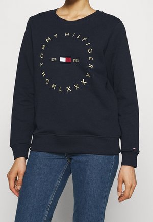 Personne portant un sweat-shirt Tommy Hilfiger bleu marine avec un texte circulaire jaune et un logo au-dessus d'un jean bleu.