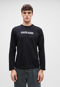 Camisola de manga longa preta feita de tecido suave, com um logotipo branco "CALVIN KLEIN" em destaque no peito. Design de gola redonda.