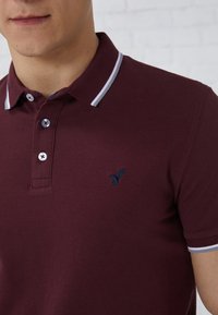 Burgunderfarbiger Poloshirt aus strukturiertem Stoff, mit einem marineblauen gestickten Logo und kontrastierenden grauen und weißen Kragenstreifen. Zwei-Knopf-Leiste.