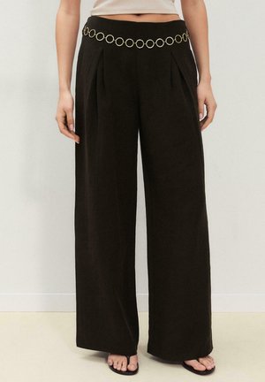 Kvinde iført sorte vidde-bukser med høj talje og læg samt en dekorativ guldkæde som bælte, kombineret med sorte sandaler.