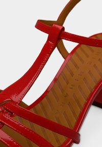 Κόκκινο σανδάλι από λουστρίνι με λουράκια και τακούνι τύπου block, με καστανή υφασμάτινη εσωτερική σόλα που φέρει ανάγλυφο το όνομα της μάρκας.
