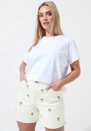 Lyshåret kvinde i hvid t-shirt og beige shorts med palmebladtryk, stående med hænderne i lommerne mod hvid baggrund.