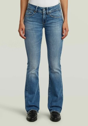 Frau trägt hellblaue Bootcut-Jeans, weißes Oberteil und schwarze Schuhe, steht mit entspannten Armen an den Seiten vor einfachem Hintergrund.