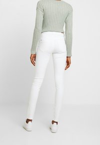 Person som bär vita slim fit jeans, vita sneakers och en ljusgrön ribbad långärmad tröja, står mot en enkel ljus bakgrund.