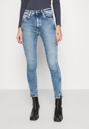 Vrouw met vervaagde blauwe skinny jeans met ritsen bij de enkels, zwarte enkellaarzen en een donker top met lange mouwen tegen een effen achtergrond.