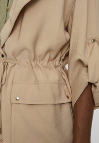 Light beige jacket with elastic waist, long sleeves, and front pockets. Features gold-tone snap details and a smooth, textured fabric.