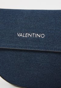 Jeansväska i mörkblå färg, med rundad form, slät textur och en metallisk "VALENTINO"-logotyp på framsidan.