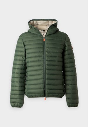 Homme debout portant une doudoune verte avec capuche, chemise marron, pantalon noir et baskets blanches sur un fond blanc uni.