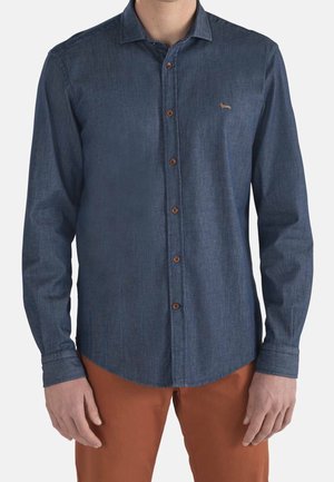 Man draagt een donkerblauw overhemd met lange mouwen, bruine knopen en een klein geborduurd logo, gecombineerd met roestkleurige broek.