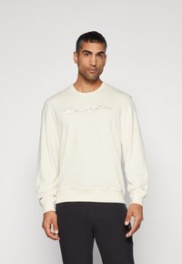 Creme sweatshirt med broderet "Champion" logo i et buet design. Har lange ærmer og ribbet krave og kant. Sorte bukser.