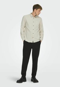Camisa abotonada beige claro con un bolsillo, de manga larga y botones marrones, combinada con pantalones rectos negros y zapatos negros.