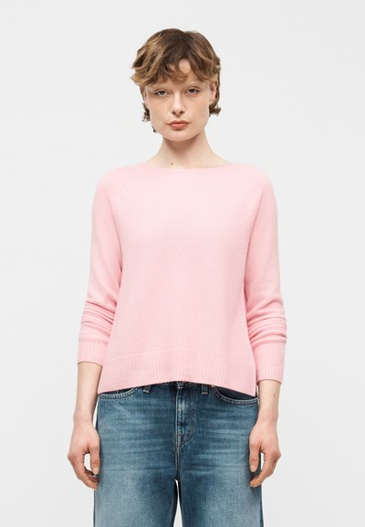 Jeune personne aux cheveux courts portant un pull à manches longues rose clair et un jean bleu en denim, debout devant un fond blanc uni.