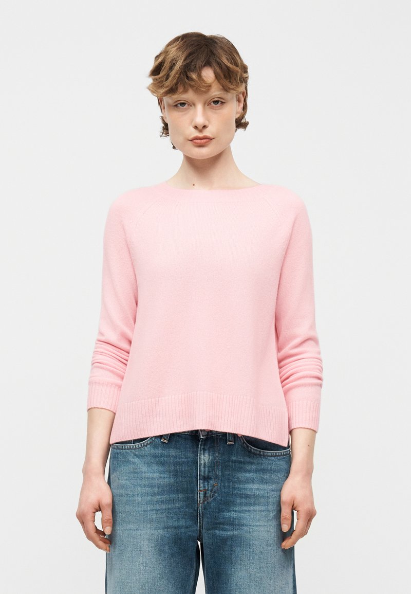 Jeune personne aux cheveux courts portant un pull à manches longues rose clair et un jean bleu en denim, debout devant un fond blanc uni.