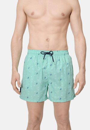 Hombre vistiendo pantalones cortos de baño verde claro con pequeños patrones de bicicletas azules y un cordón azul marino, de pie frente a un fondo liso.