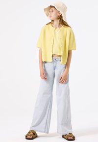 Cárdigan de punto amarillo de manga corta sobre un top corto a juego, combinado con unos pantalones vaqueros de pierna ancha en azul claro y sandalias marrones. Sombrero tipo bucket en color crema incluido.