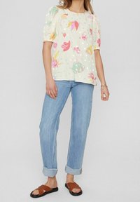 Ljusgul blus med blommönster och prickigt mönster, korta puffärmar, kombinerad med ljusblå uppkavlade jeans och bruna slide-sandaler.