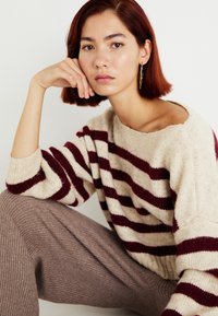 Gestreepte trui in crème en bordeaux, gemaakt van zachte brei. Geribbelde manchetten. Model draagt een aansluitende beige geribbelde broek, met een neutrale achtergrond.