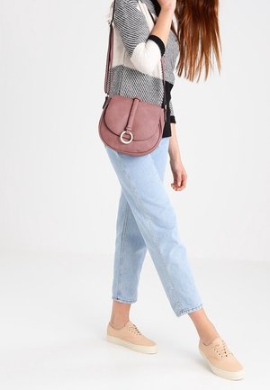 Torebka crossbody w mauve skórze z okrągłym metalowym detalem, zestawiona z jasnoniebieskimi dżinsami i beżowymi trampkami. Posiada pasek na ramię.