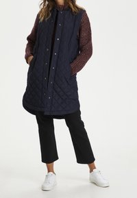 Gilet imbottito navy con bottoni a pressione, tasche laterali e orlo arrotondato, indossato sopra un maglione in maglia bordeaux e abbinato a pantaloni neri e sneakers bianche.